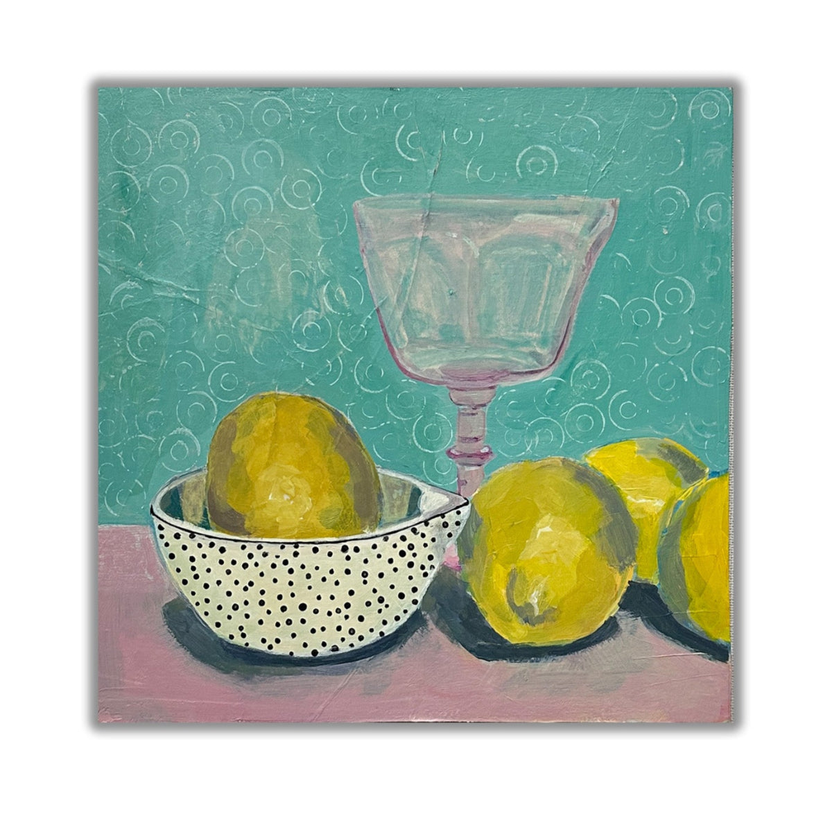 group of lemons on a pink table with a turqoise background. pink wine glass and a polka dot bowl holding another lemon.