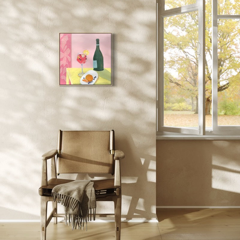 A framed square wall art print depicting a still life with a green bottle, pink flowers, and a yellow table, hung on a beige wall in a sunlit room above a wooden armchair.