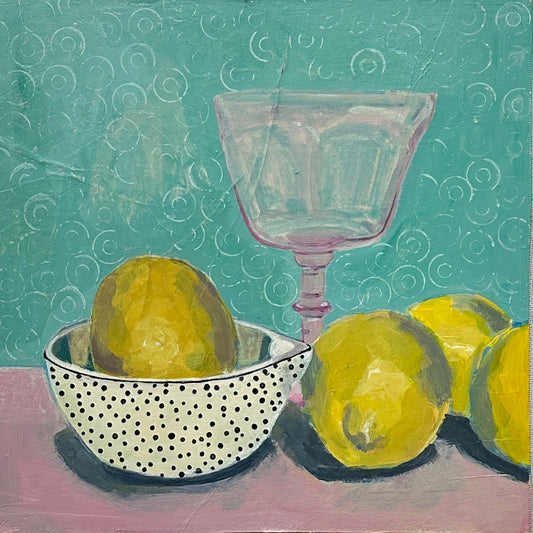 group of lemons on a pink table with a turqoise background. pink wine glass and a polka dot bowl holding another lemon. 