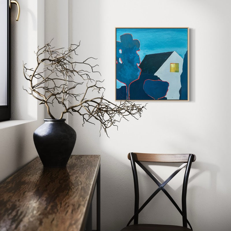 A framed abstract painting featuring a white house against  blue sky, hanging on a white wall above a wooden chair and next to a dark vase with dried branches.
