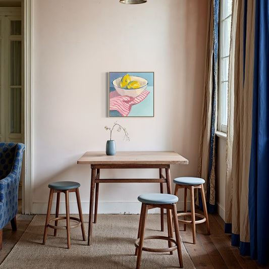 still life painting with white bowl of lemons hanging on pale pink wall above a wood table with stools. 