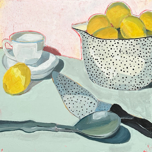 still life of polka dot bowl holding lemons, a white teacup with a lemon leaning on it, and kitchenware, sitting on a mint table with a pink background.