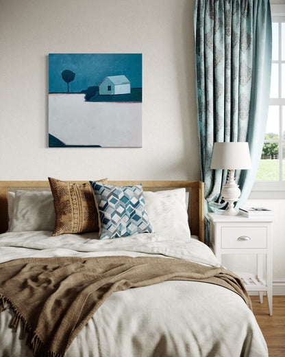 Painting of a solitary house painted in blues and pinks styled in a bedroom with cream comforter and brown throw blanket.