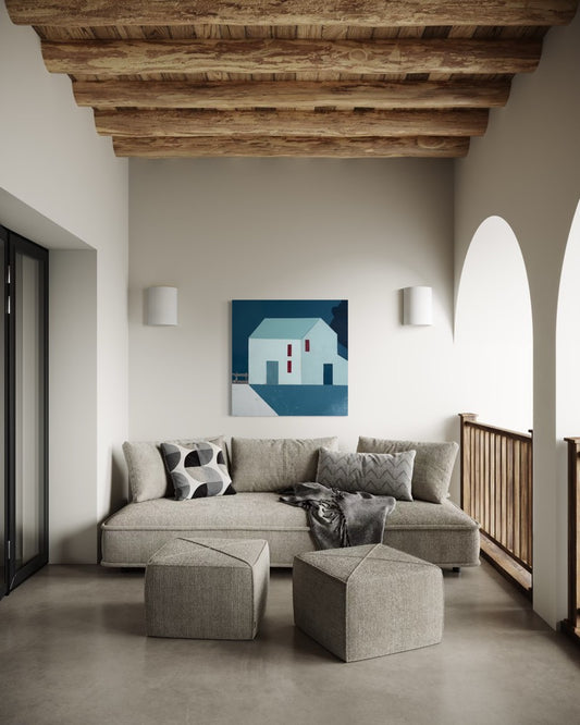 A modern living room interior featuring a light gray sectional sofa with matching geometric ottomans, a framed abstract house painting on the wall, exposed wooden ceiling beams, and concrete floors.