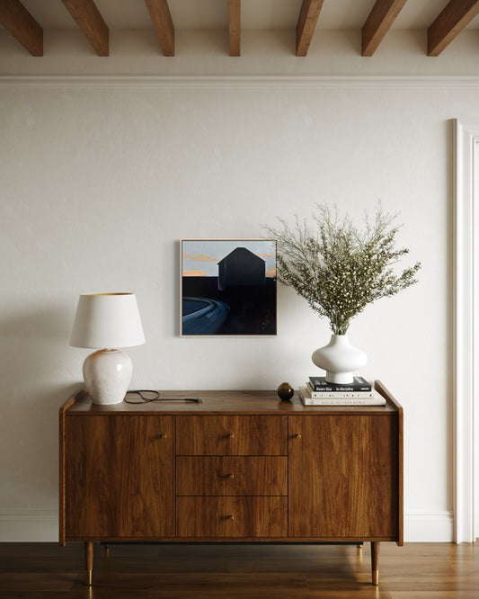 A mid-century modern wooden sideboard cabinet styled in a minimalist room, topped with a white ceramic table lamp, a small framed print, books, and a white vase holding dried branches.