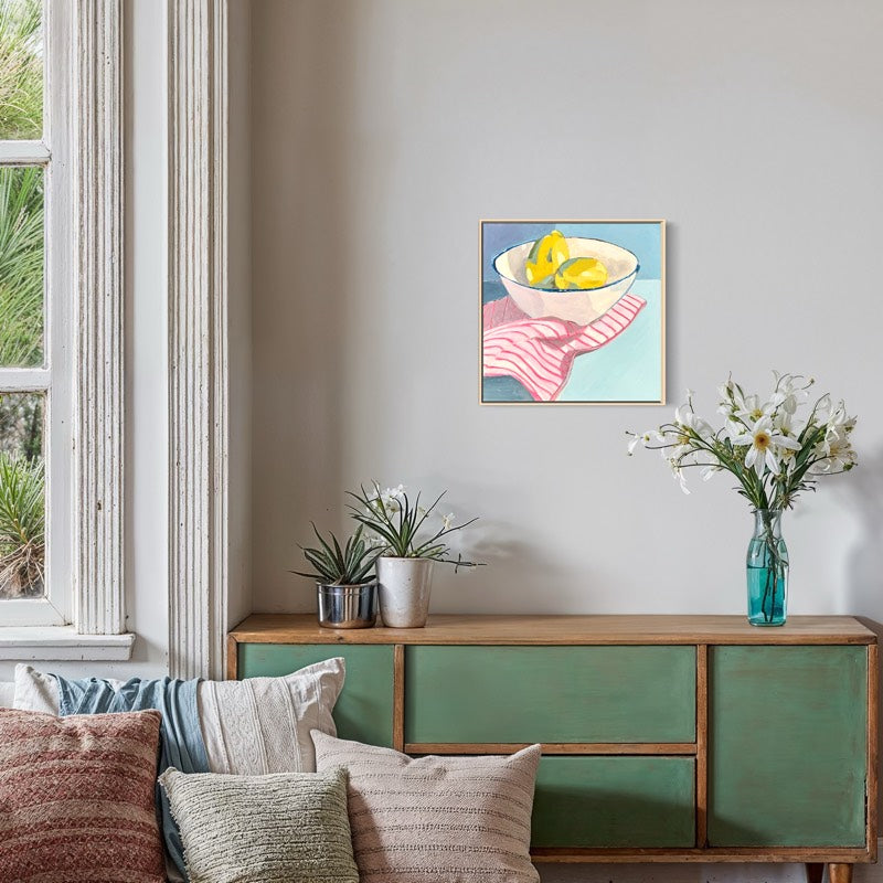 A framed art print depicting a still life of lemons in a bowl with a pink striped cloth, hanging on a beige wall above a green wooden sideboard in a styled room.
