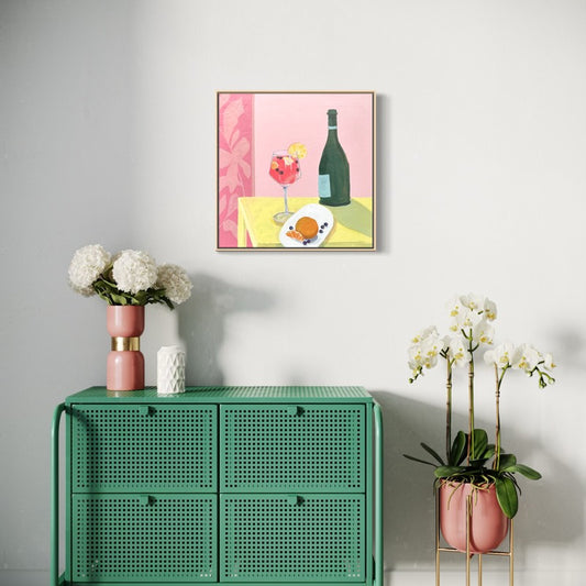 A framed art print depicting a pink cocktail, a wine bottle, and a plate of food on a yellow table, hung on a light gray wall above a green cabinet with pink and white floral arrangements.